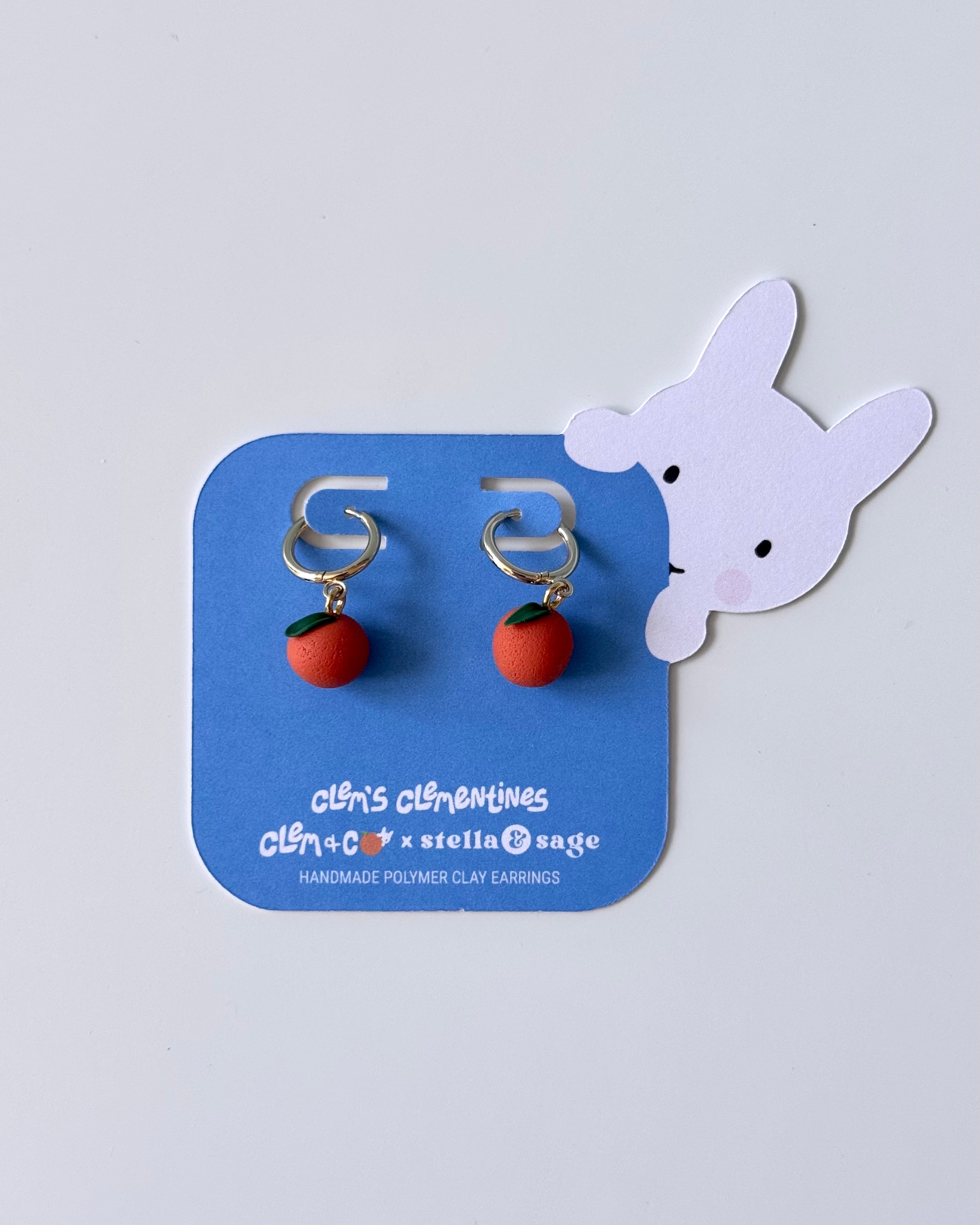 Pair of clementine shaped earrings on a blue card with a white bunny illustration peering around the side.