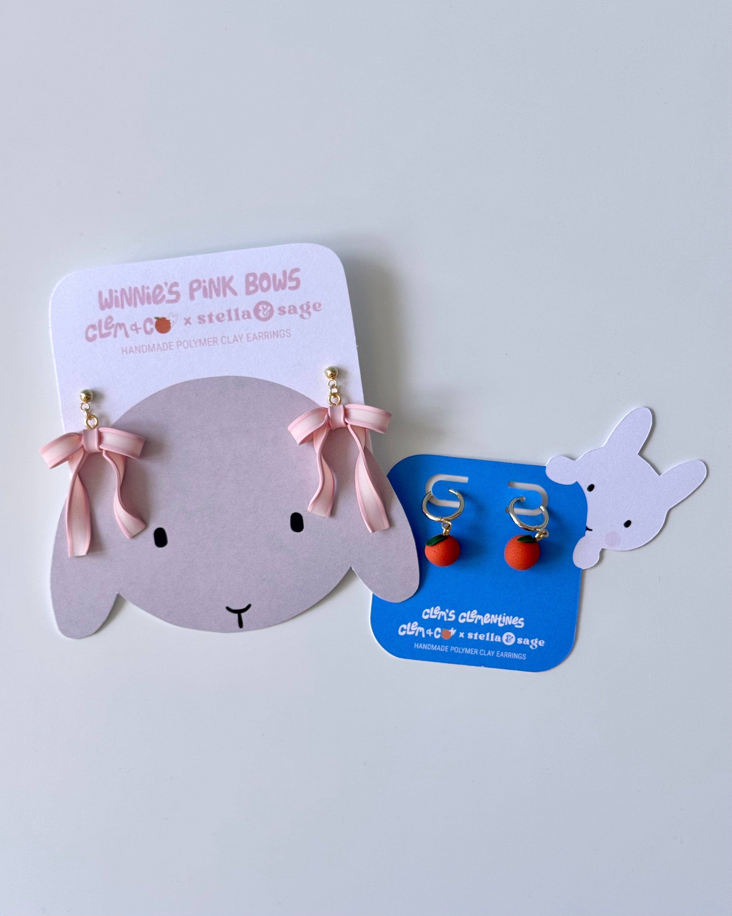Two sets of earrings on backing cards. The first is a set of pink bows and the second is a set of clementines.