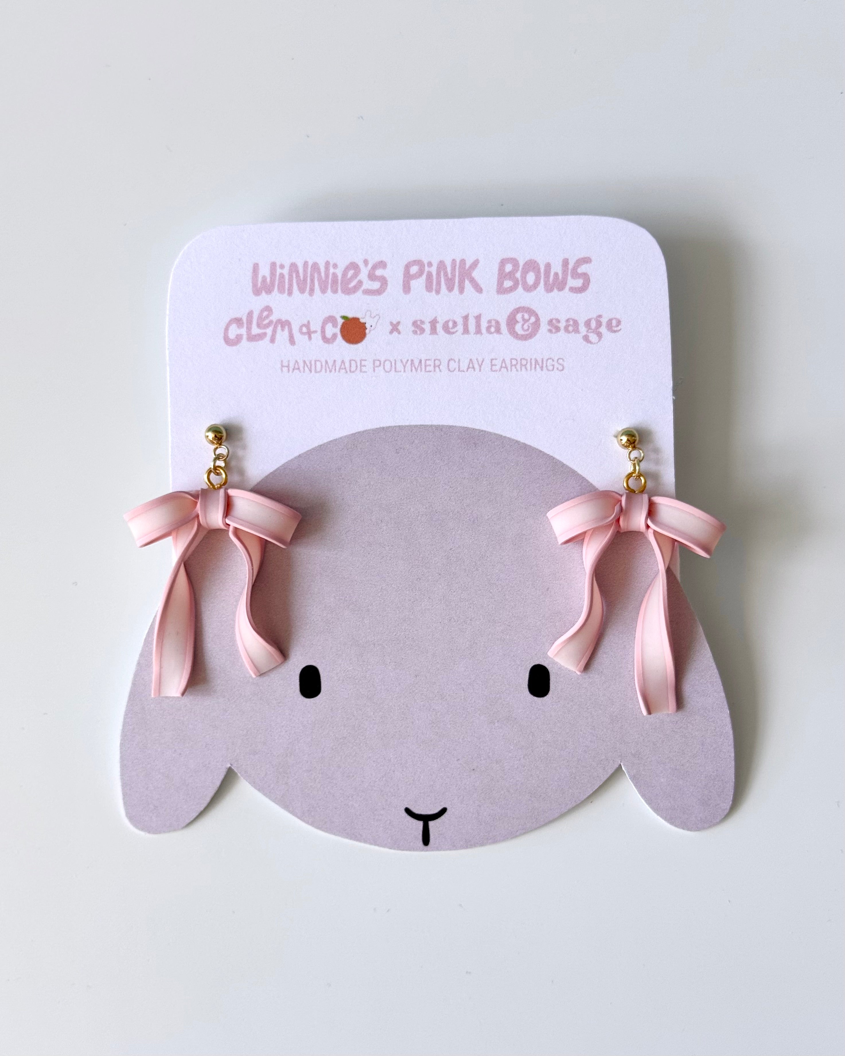 Two polymer clay pink bow dangle earrings on a backing card with an illustration of a gray bunny on it.