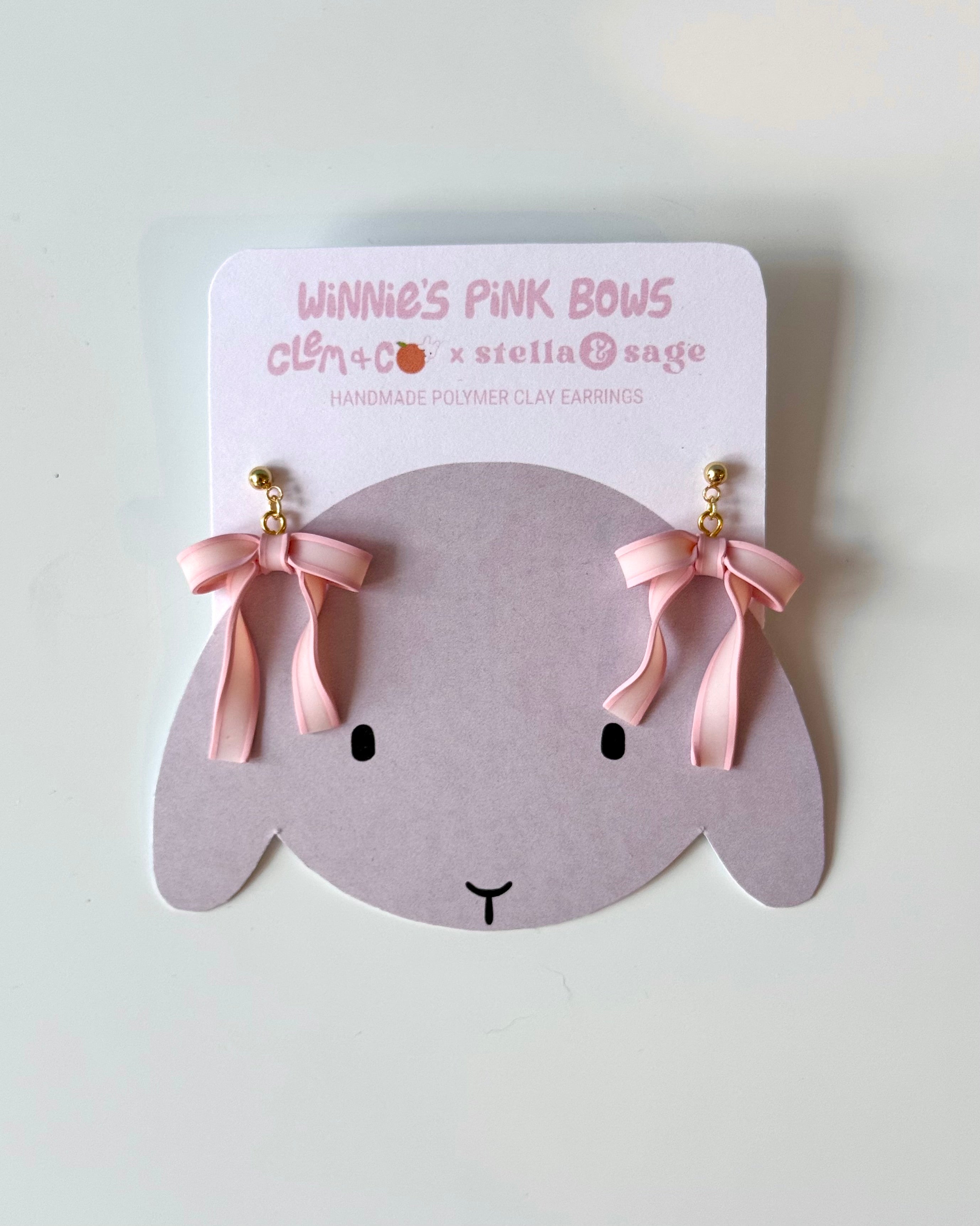 Two polymer clay pink bow dangle earrings on a backing card with an illustration of a gray bunny on it.